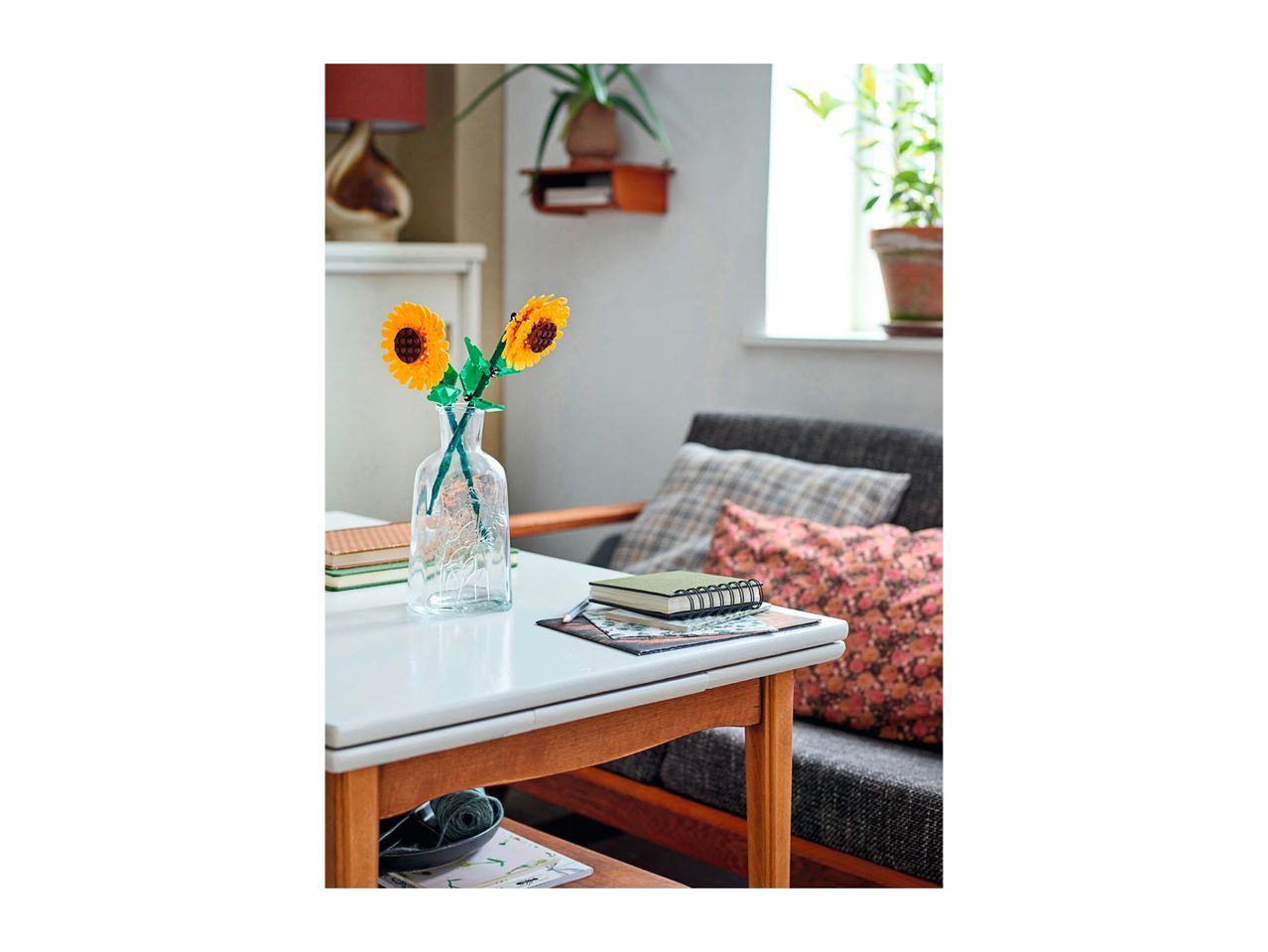 Lego sunflowers in a vase on a white table with notebooks and a patterned cushion.