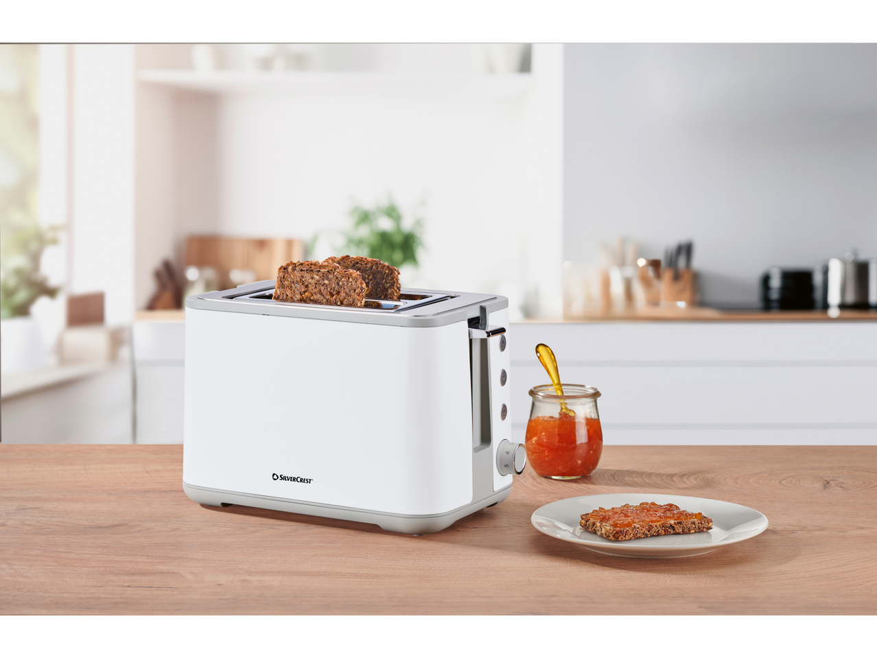 White toaster with two slices of seeded bread, a jar of jam, and a jam-covered slice on a plate.