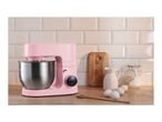 Pink stand mixer with a stainless steel bowl, eggs, flour, and a wooden spoon on a kitchen counter.