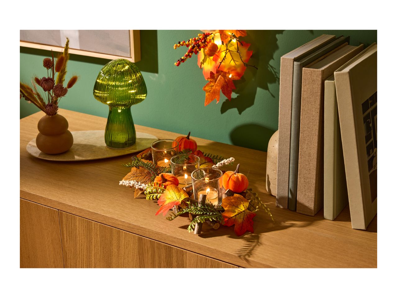 Autumnal home decor with a green mushroom lamp, candles, pumpkins, and books on a wooden cabinet.