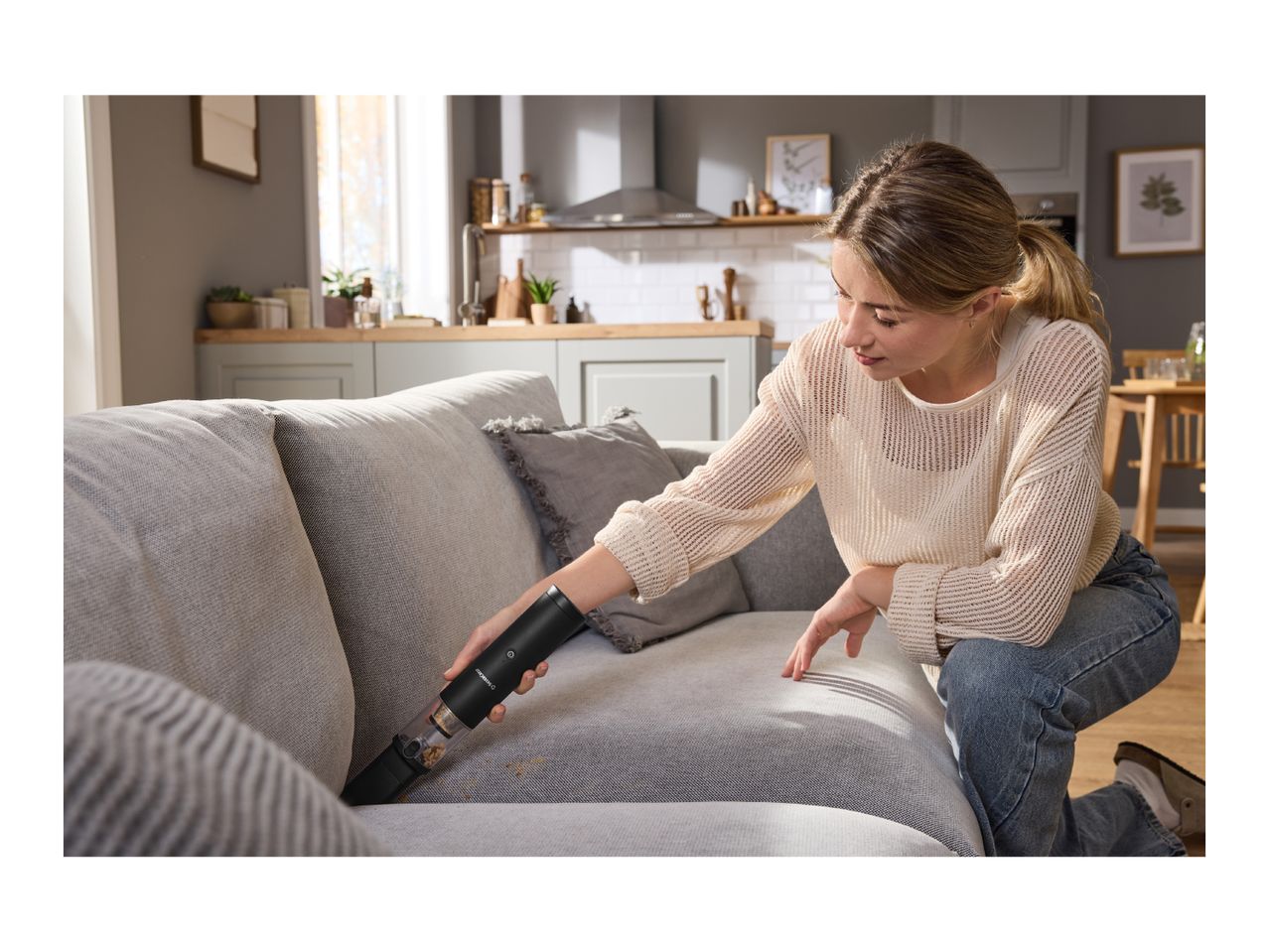 Woman cleaning a grey sofa with a black handheld vacuum cleaner in a modern living room.