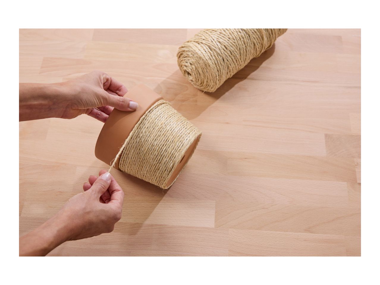 Hands wrapping a terracotta pot with natural twine on a wooden surface.