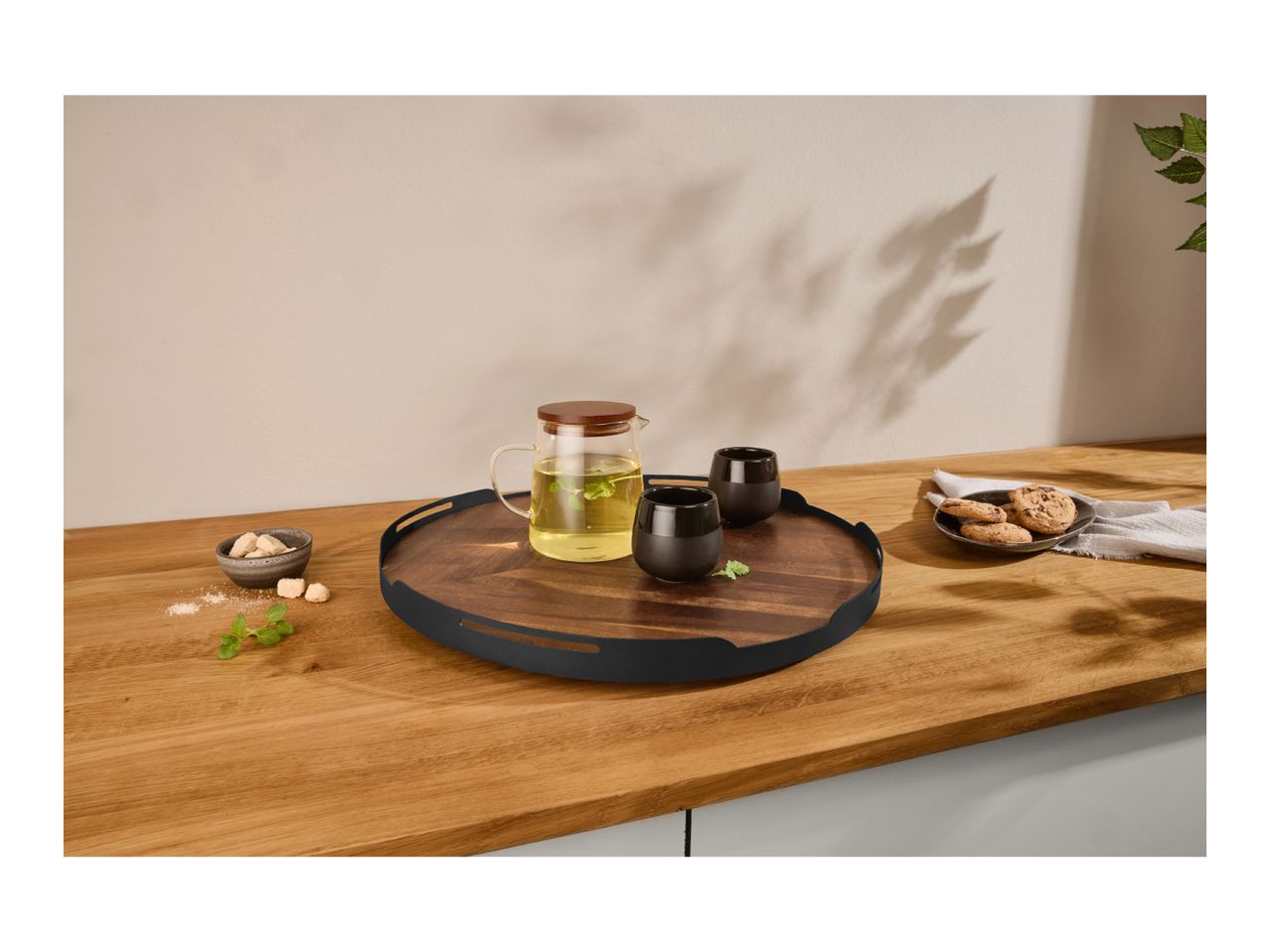 Wooden serving tray with a glass teapot, two black cups, sugar cubes, and cookies.
