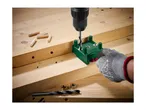 Drilling holes in wood with a drill and a green dowel jig, with wooden dowels nearby.
