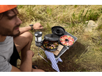 Man camping with a portable grill, camping lantern, and fresh food on a table.
