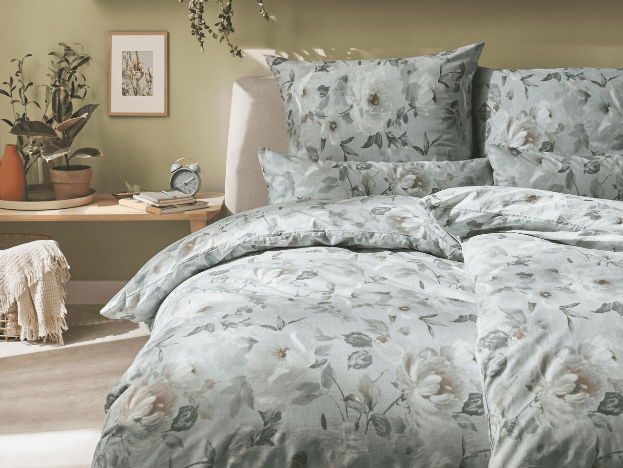 Ein Bett mit floraler Bettwäsche in einem Schlafzimmer mit Pflanzen und einem Wecker auf einem Nachttisch.