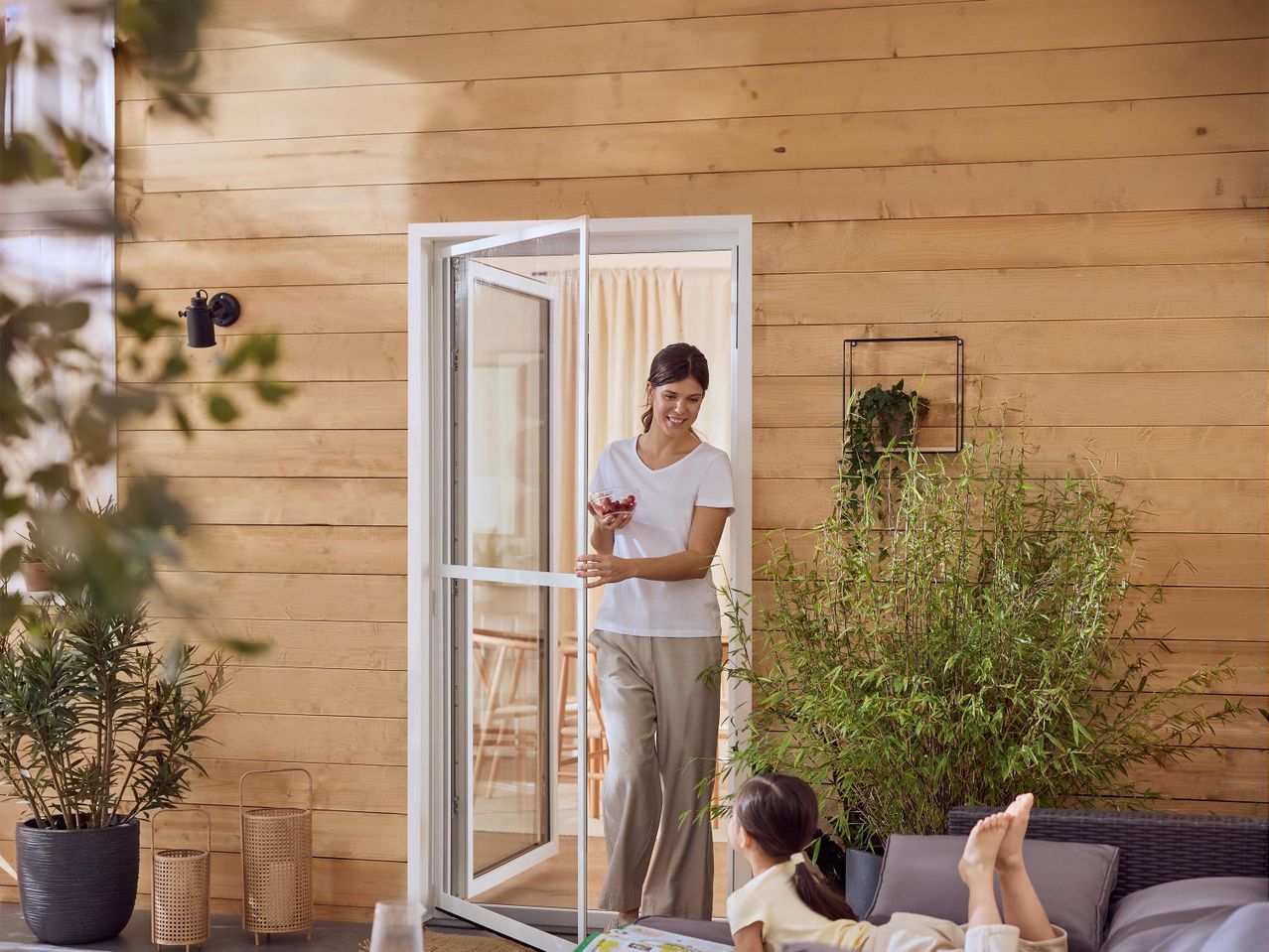 Nő nyitott teraszajtóval, kezében tál cseresznyével, miközben egy gyermek fekszik a kanapén.