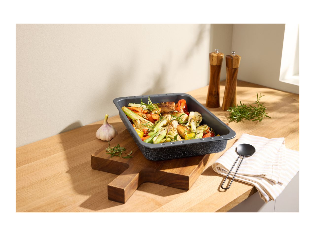 Roasting pan with roasted vegetables, garlic, rosemary, and wooden salt and pepper mills.
