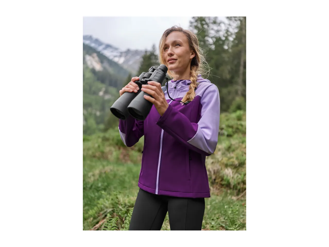 Woman in a purple outdoor jacket holding binoculars, looking at mountains.