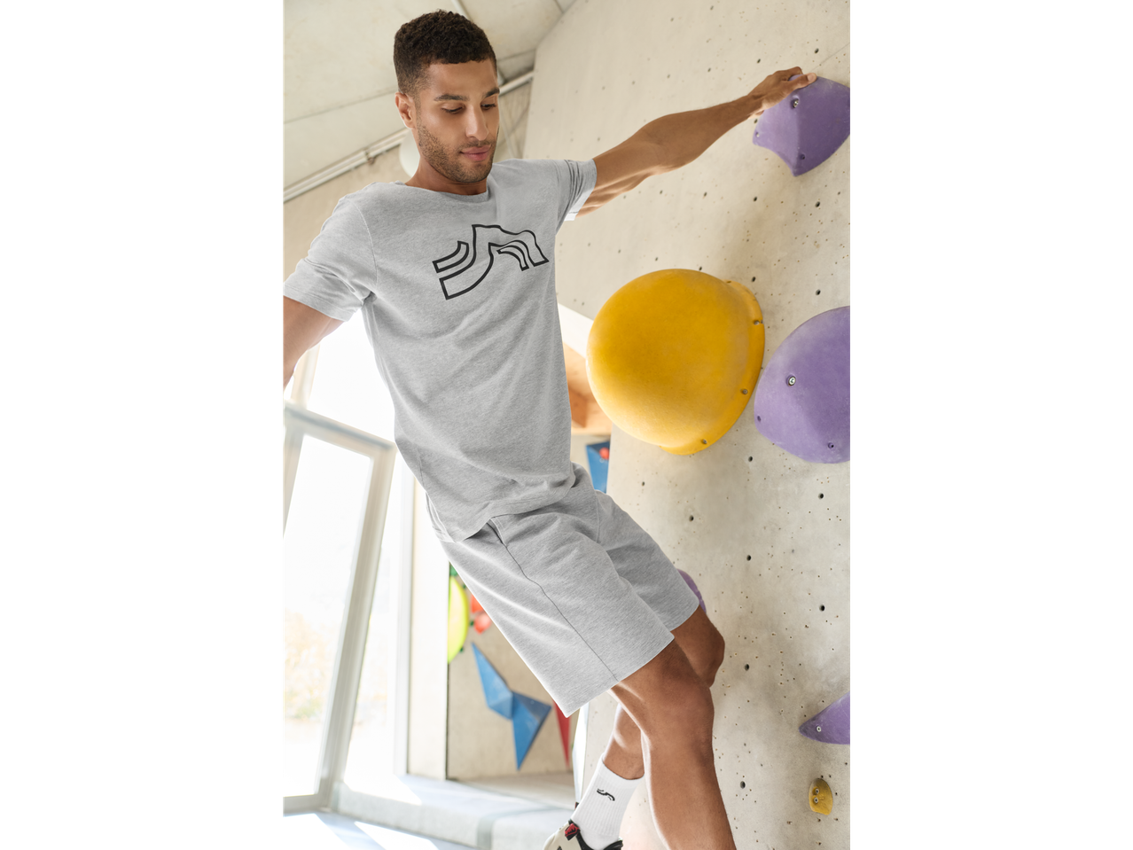 Muškarac u sivoj majici i šortsu penje se po zidu za penjanje, noseći bele Crivit sportske čarape.