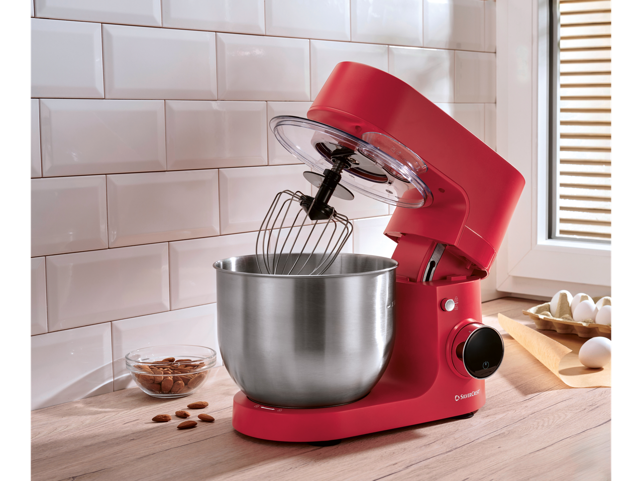 Robot de bucătărie roșu SilverCrest cu tel, bol, migdale și ouă pe un blat de lemn.