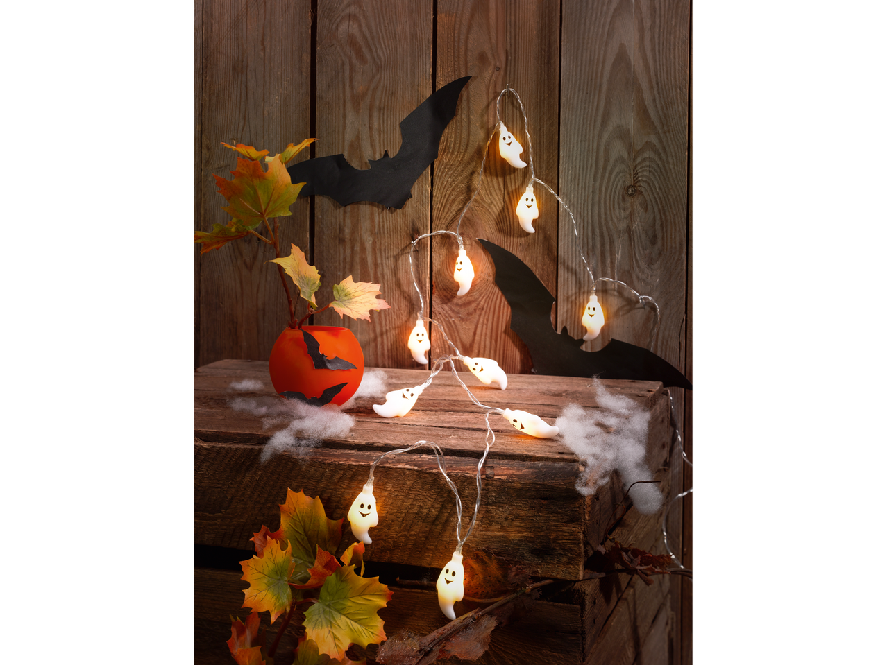 Decor de Halloween cu luminițe fantomă, lilieci de hârtie și frunze de toamnă pe o ladă de lemn.