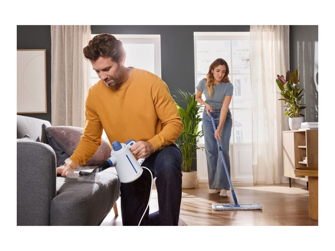 Man steam cleaning a sofa, woman mopping a wooden floor in a living room.