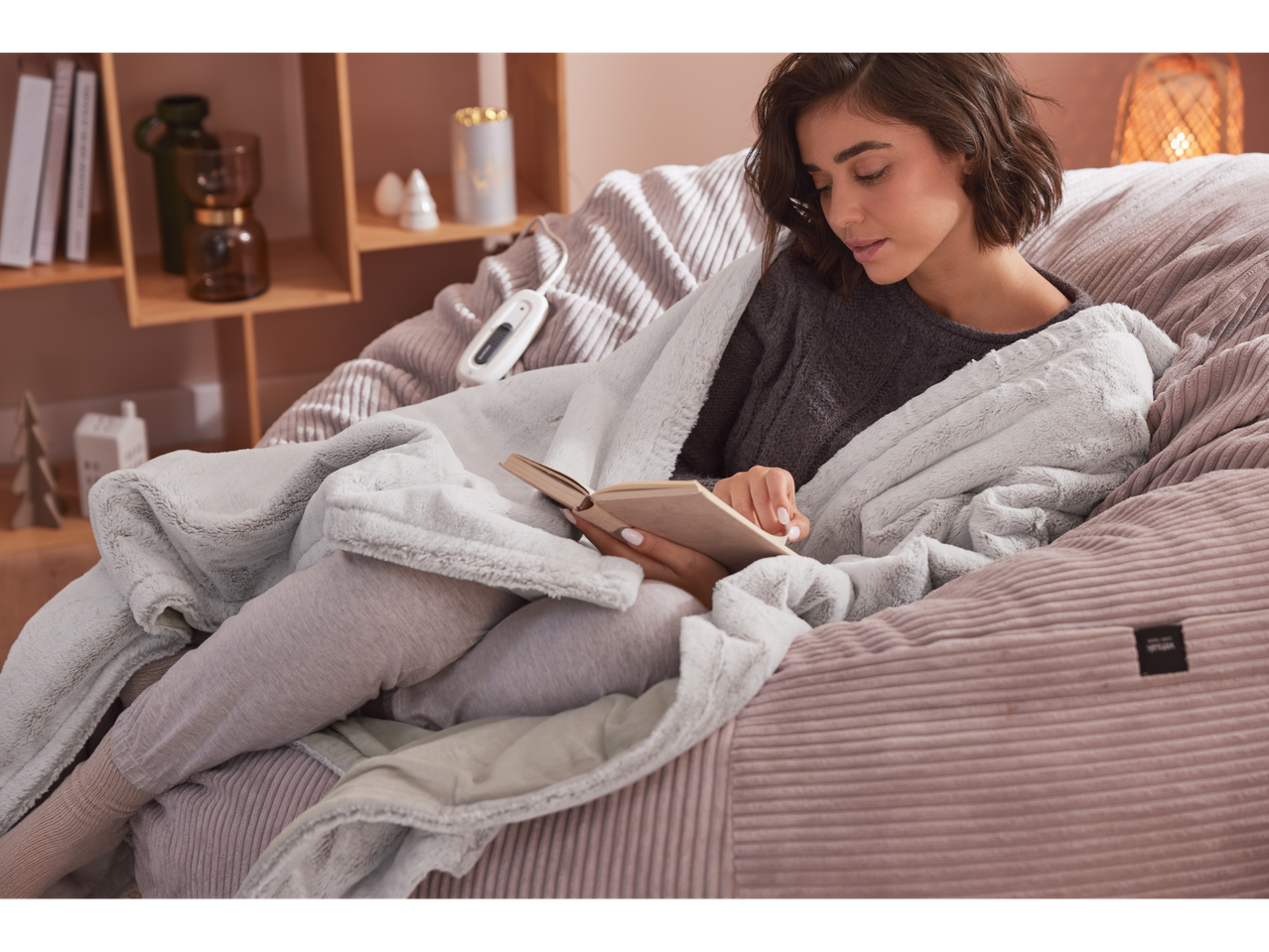Femme lisant un livre, enveloppée dans un plaid chauffant Silvercrest® sur un fauteuil beige.