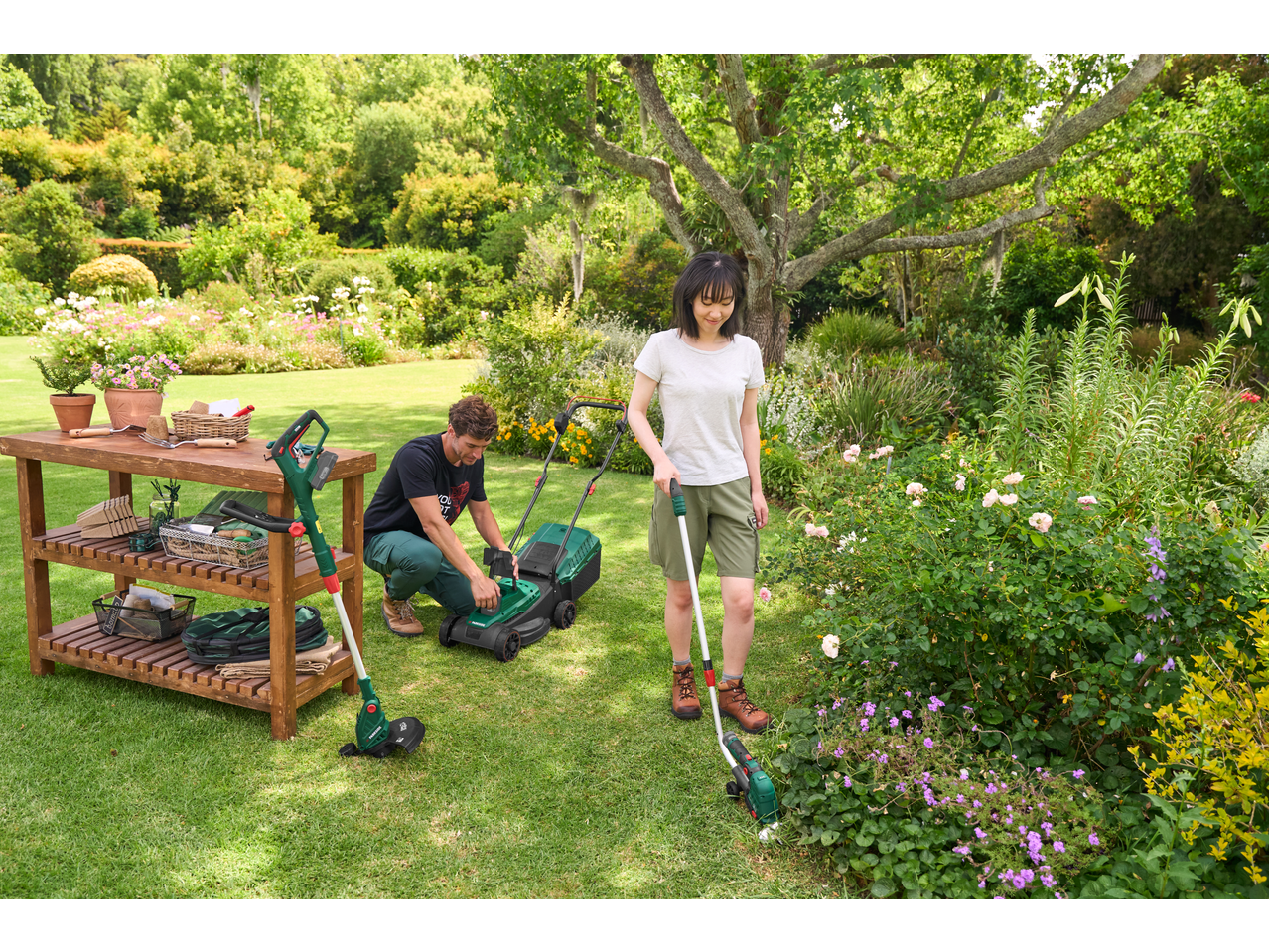 A man and a woman gardening with a lawnmower and a grass trimmer in a lush garden.