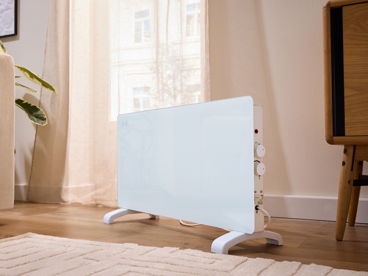 Бял конвектор Tronic® на дървен под, до прозорец с прозрачни завеси.