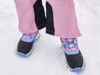 Close-up of a person wearing pink snow pants and black and purple winter boots in snow.