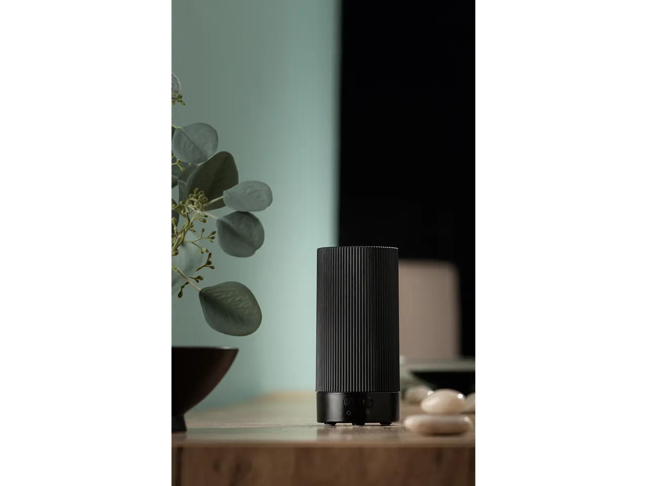 A black essential oil diffuser on a wooden surface with a plant and stones.