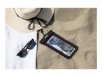 Waterproof phone pouch with a smartphone on sand next to a hat, towel, and sunglasses.