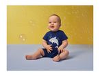 Happy baby in a blue shark-themed romper, sitting amidst bubbles on a yellow background.