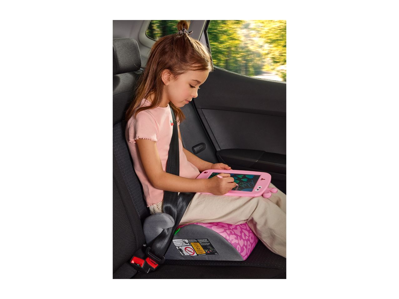 Girl in car seat drawing on a pink digital tablet, with trees visible outside.