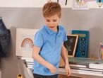 A boy in a light blue polo shirt stands at a desk with school supplies.