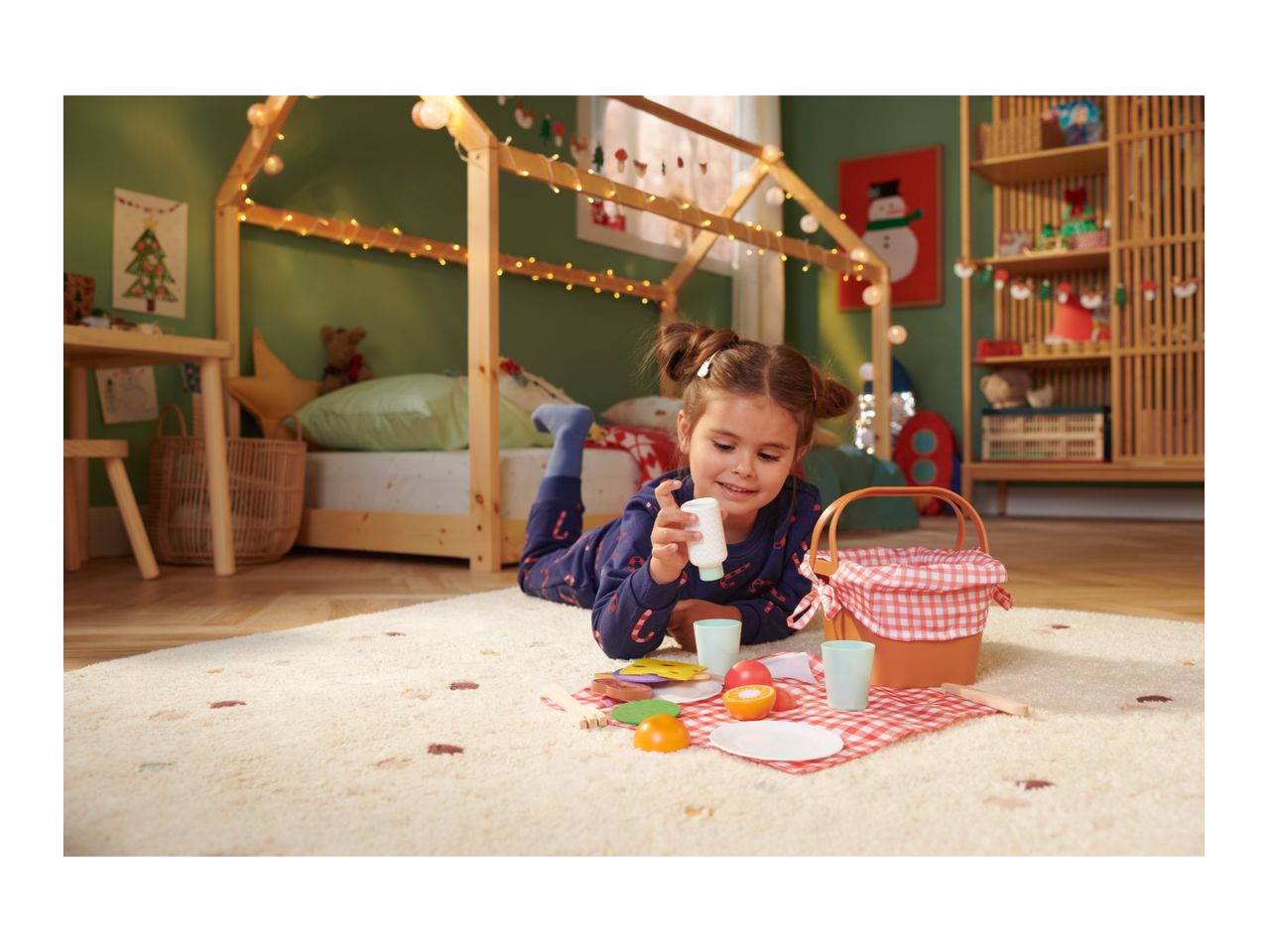 Girl playing with a toy picnic basket and food on a rug in a Christmas-themed room.