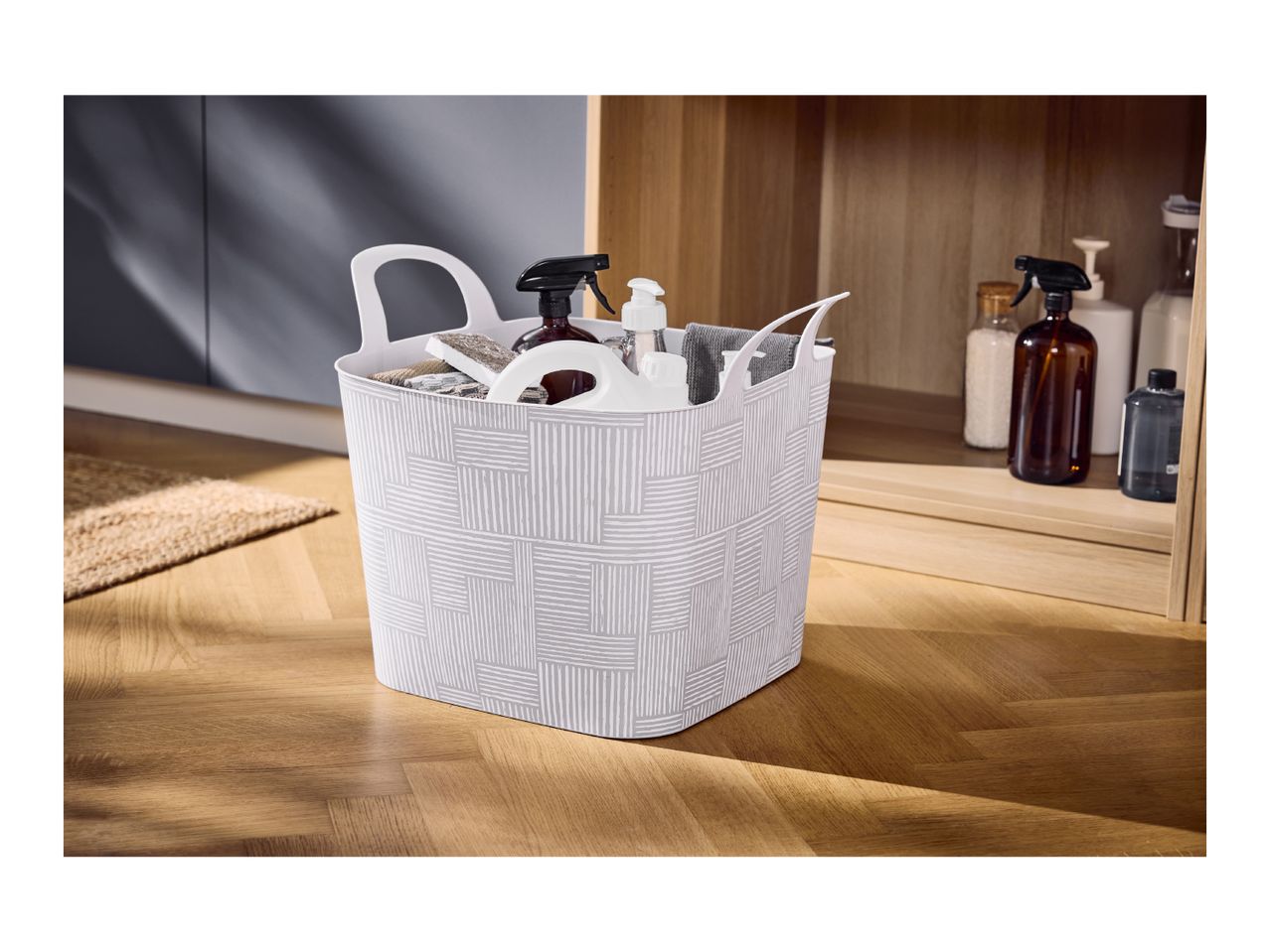 A patterned plastic storage basket filled with cleaning supplies on a wooden floor.