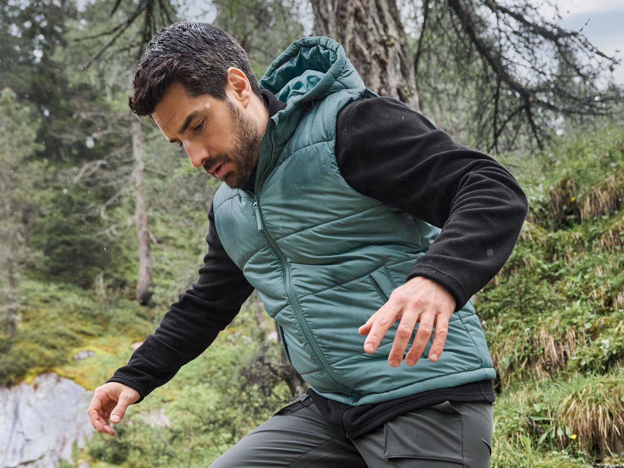 Man in a teal puffer vest and black long-sleeved shirt hiking in a forest.