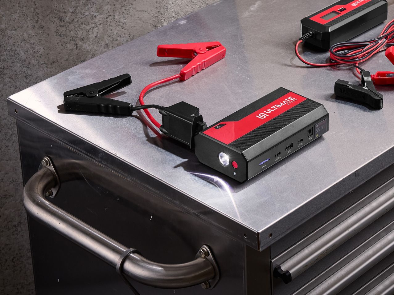 Jump starter with clamps and a battery charger on a metal workbench.