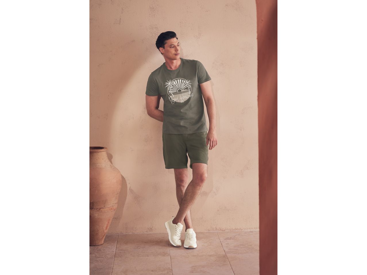 Un uomo indossa una t-shirt verde oliva con stampa a tema tropicale e pantaloncini corti, abbinati a scarpe da ginnastica bianche.