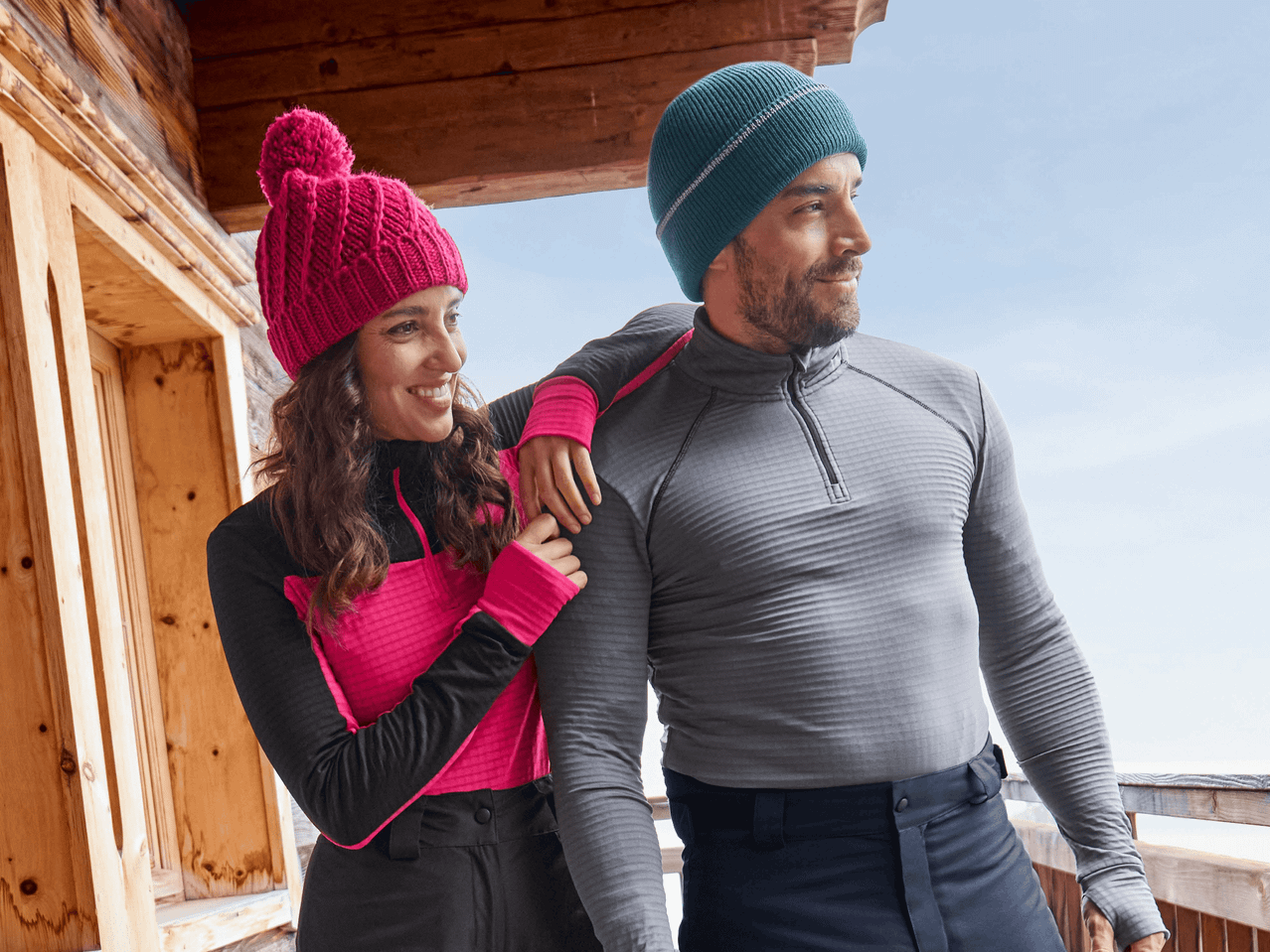 Ein Paar in Winterkleidung, die Frau trägt eine pinke Mütze und der Mann eine blaue Mütze, auf einem Balkon.