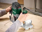 Person using a green and grey router on a wooden surface, creating a curved cut.