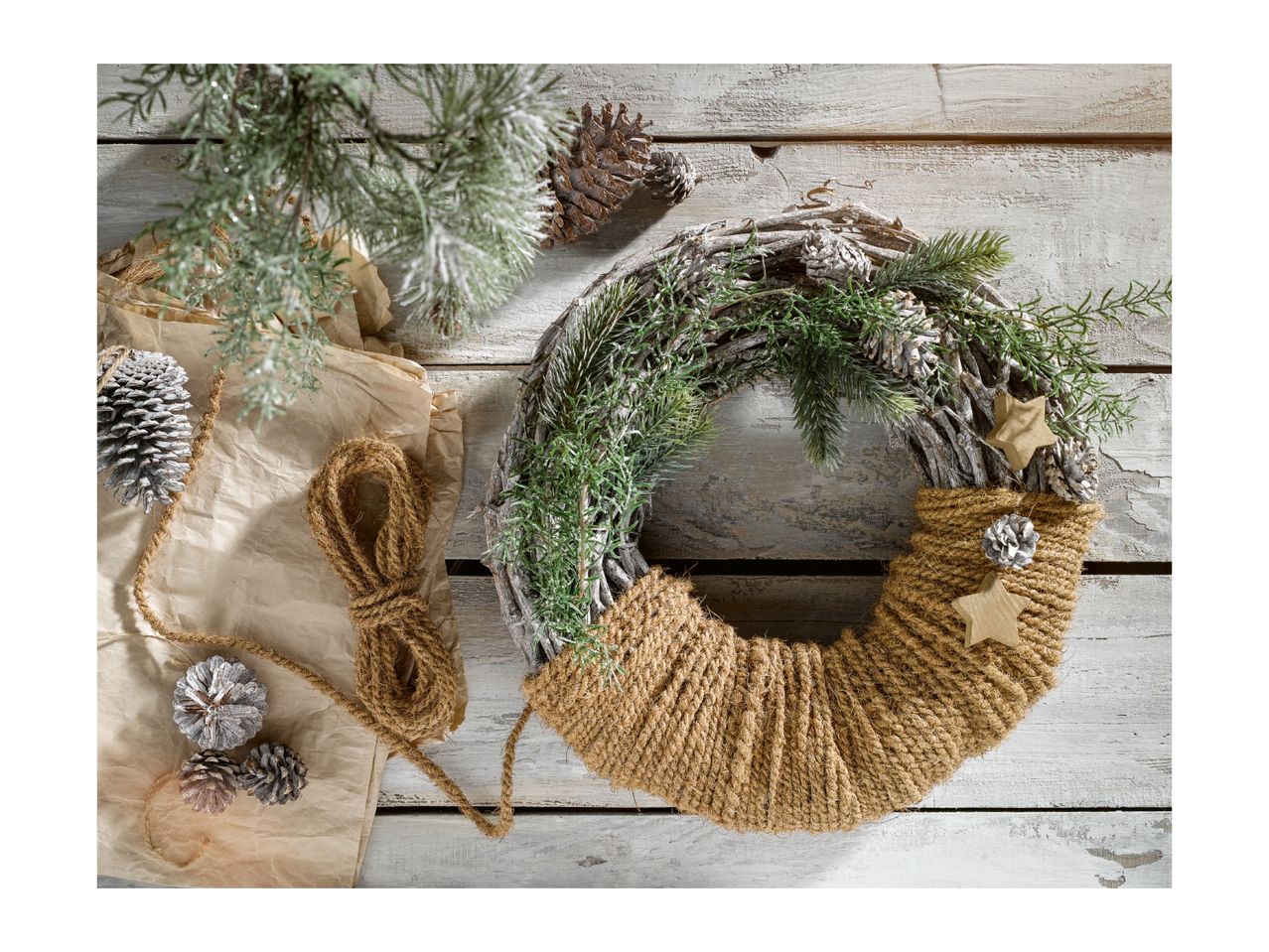 Handmade Christmas wreath with pine branches, cones, and twine on a rustic wooden background.