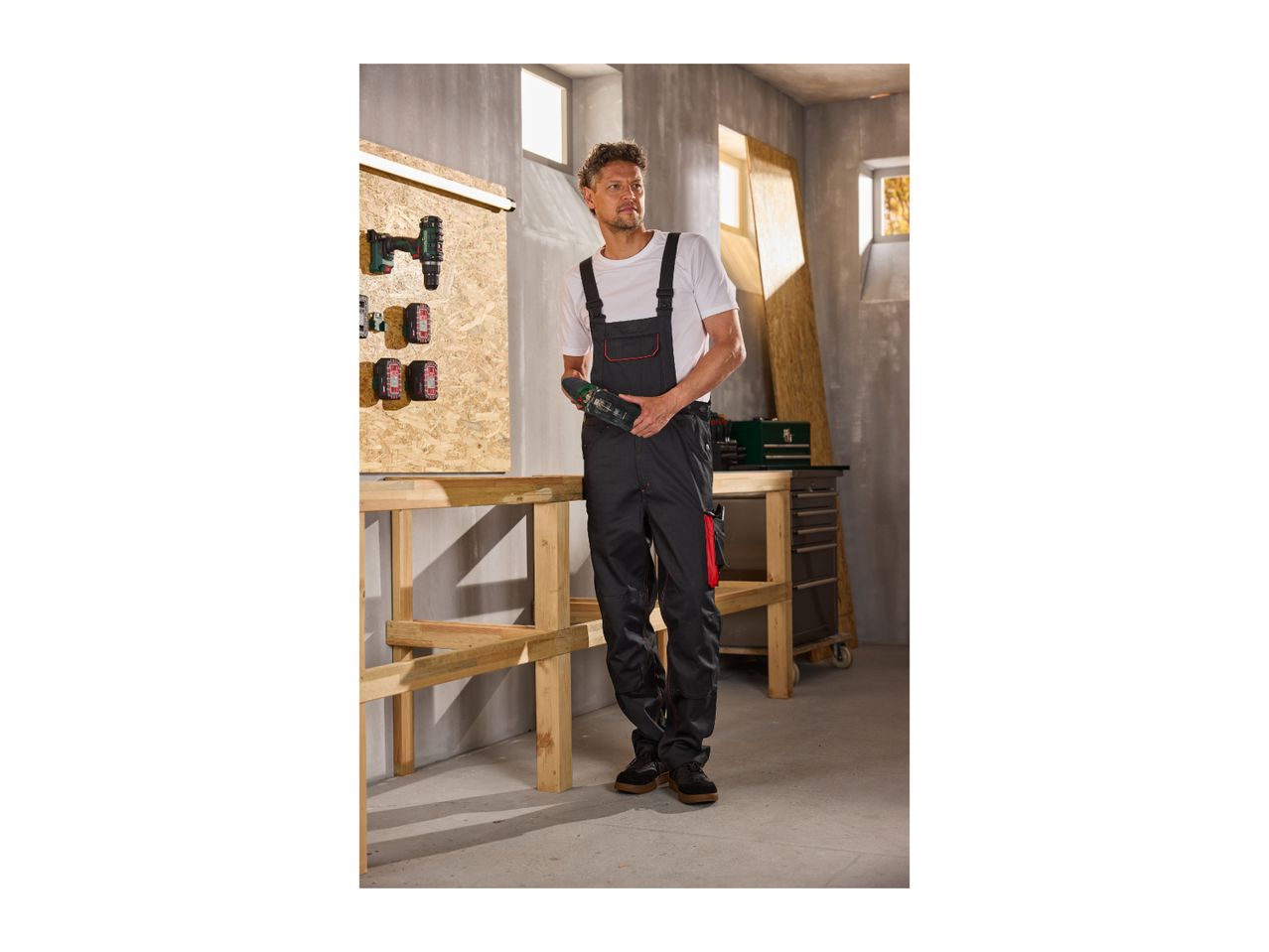 Man in work overalls holding a power tool in a workshop with tools on a wooden board.