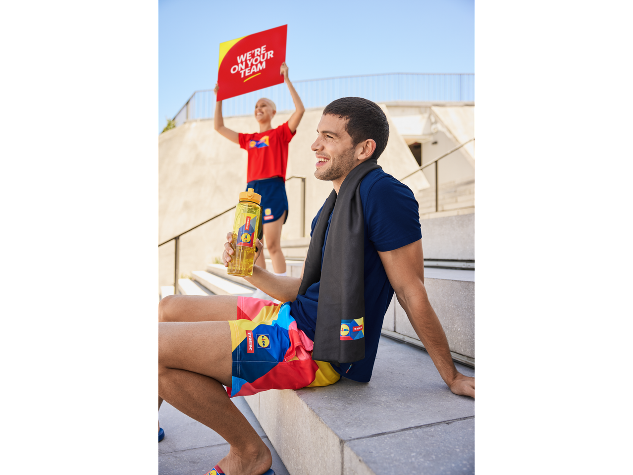 Homem sentado em degraus com uma garrafa de água Lidl Trek e uma toalha, enquanto uma mulher segura um cartaz 'Estamos na sua equipe'.