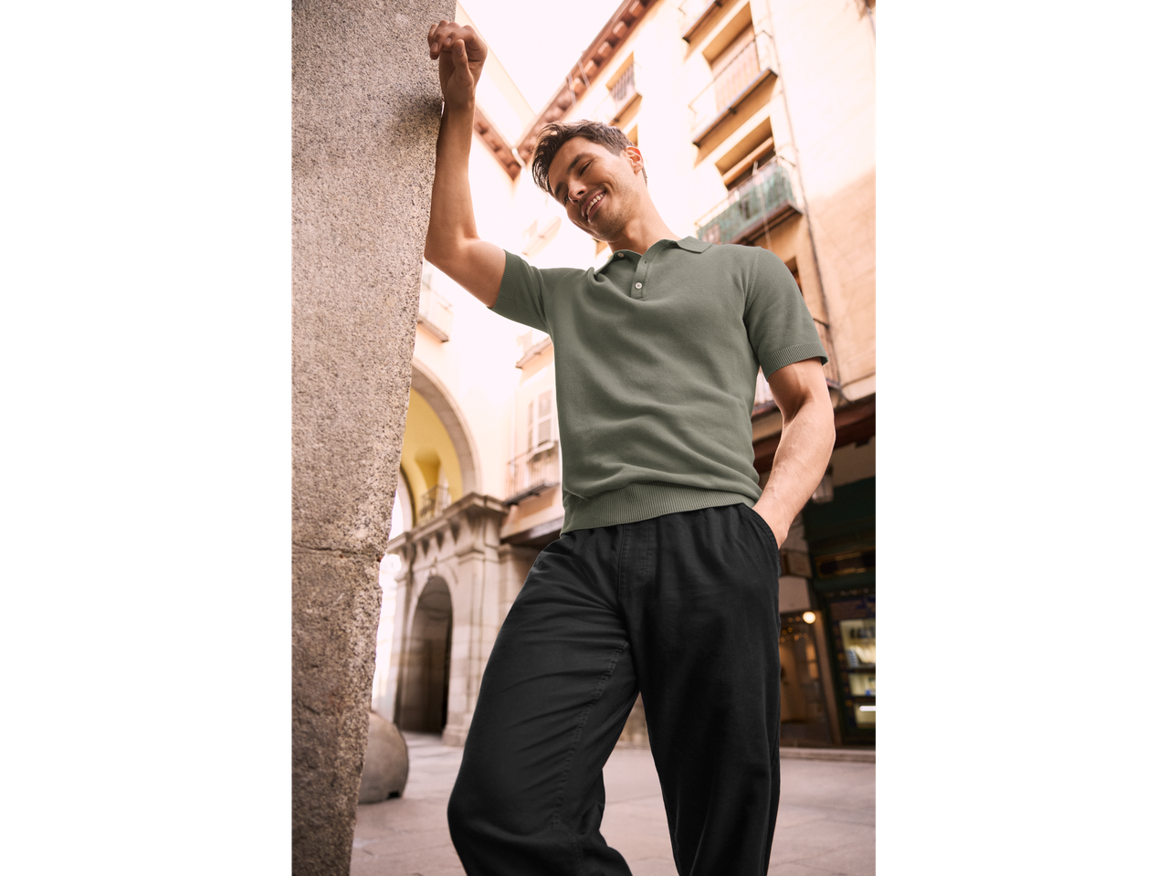 Modelo masculino vestindo um polo verde-oliva e calças pretas em uma rua europeia.
