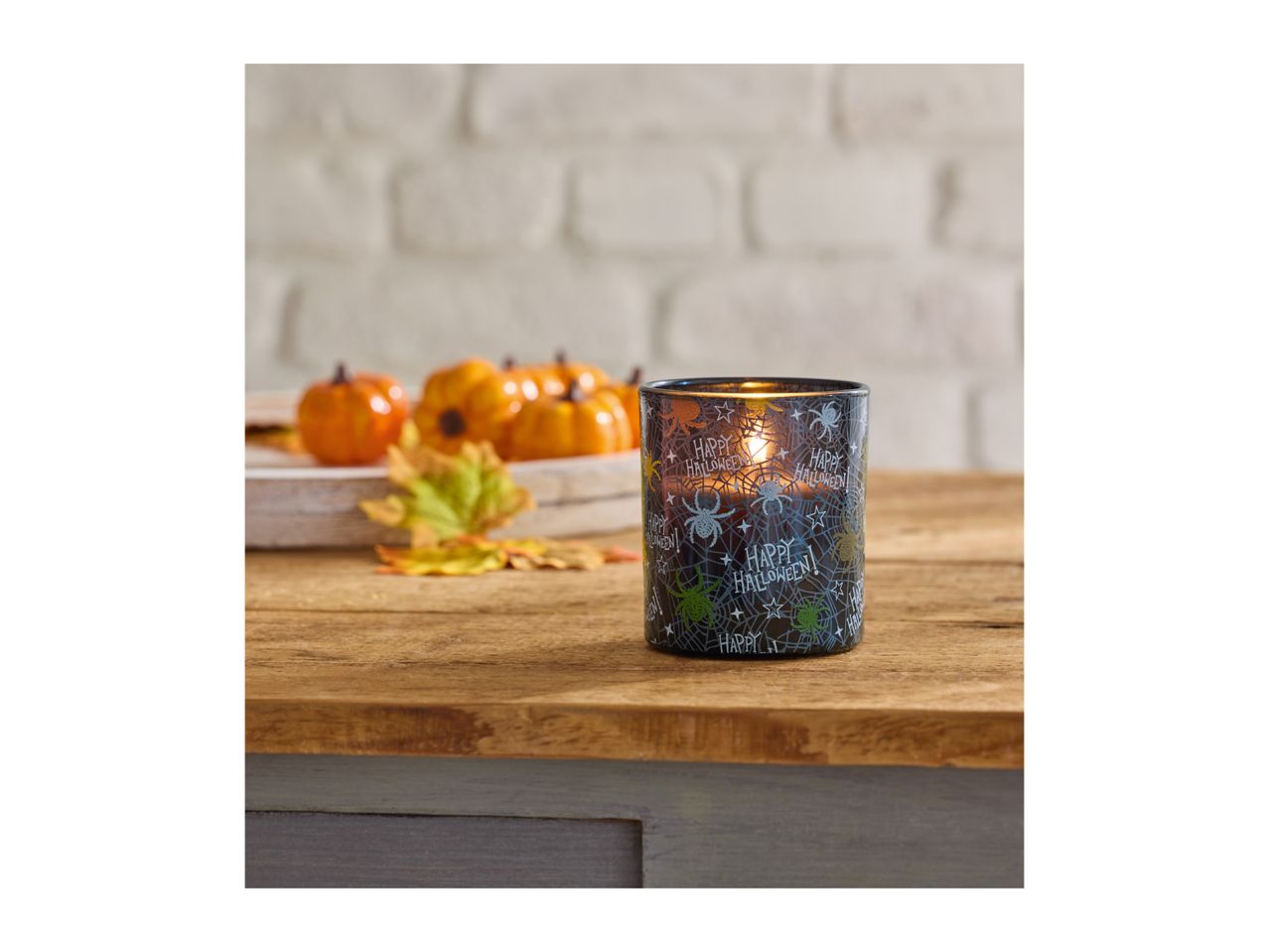 Halloween candle with spiders and text, surrounded by mini pumpkins and autumn leaves.
