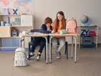 Two children at a desk, one with a Crelando pencil case, a pink backpack, and a white backpack.