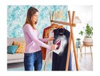 Woman steaming clothes on a wooden rack in a living room with tropical wallpaper.