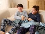 Two children in plaid pajamas and long-sleeved shirts sharing a blue blanket on a sofa.