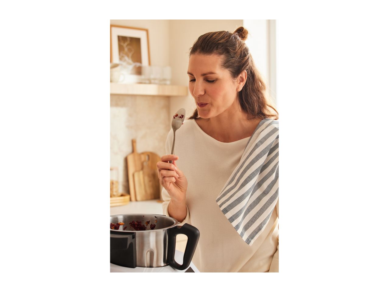 A woman tastes food from a pot with a spoon in a kitchen.
