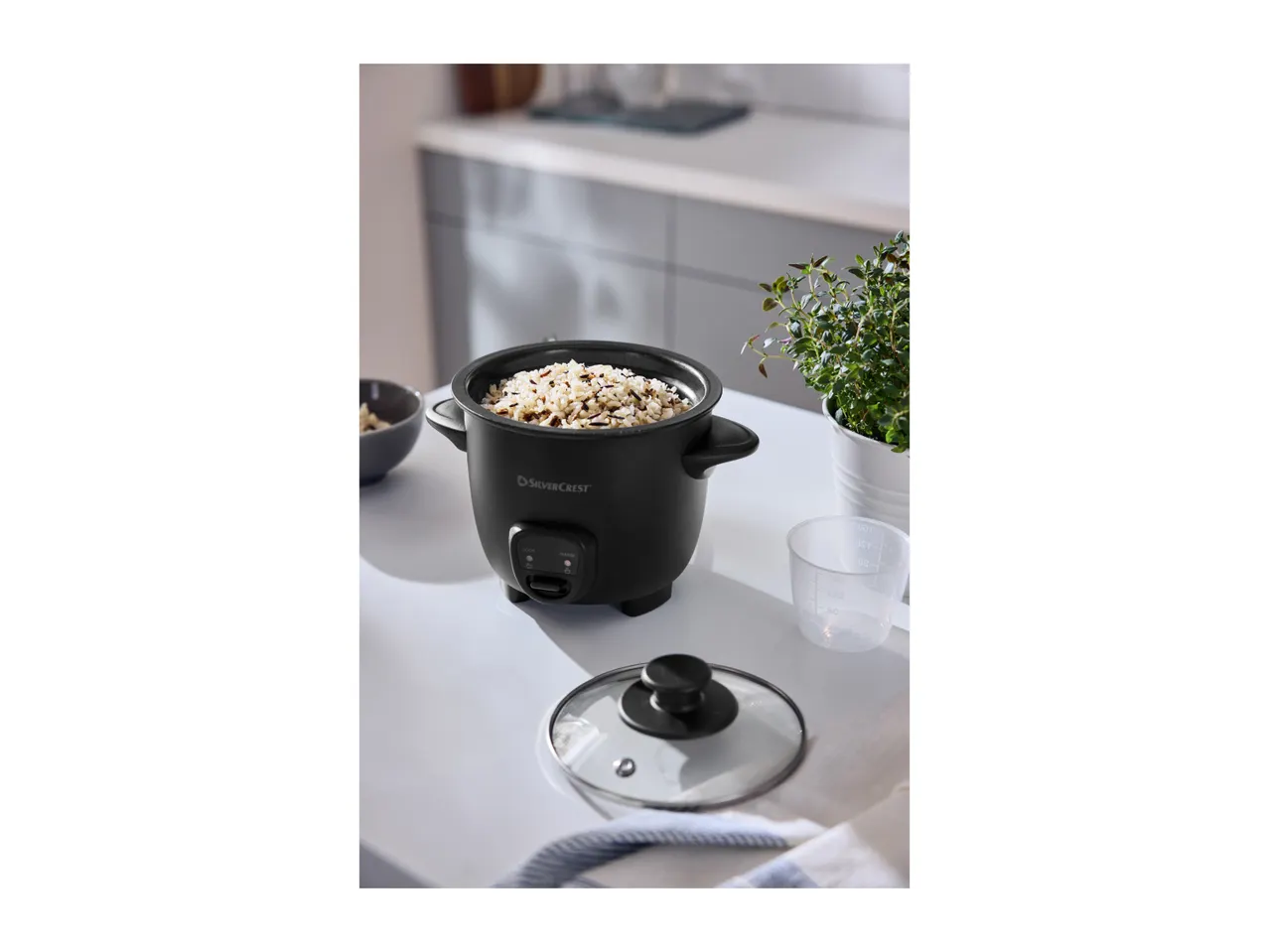 Black rice cooker filled with rice, with a measuring cup and plant on a white counter.