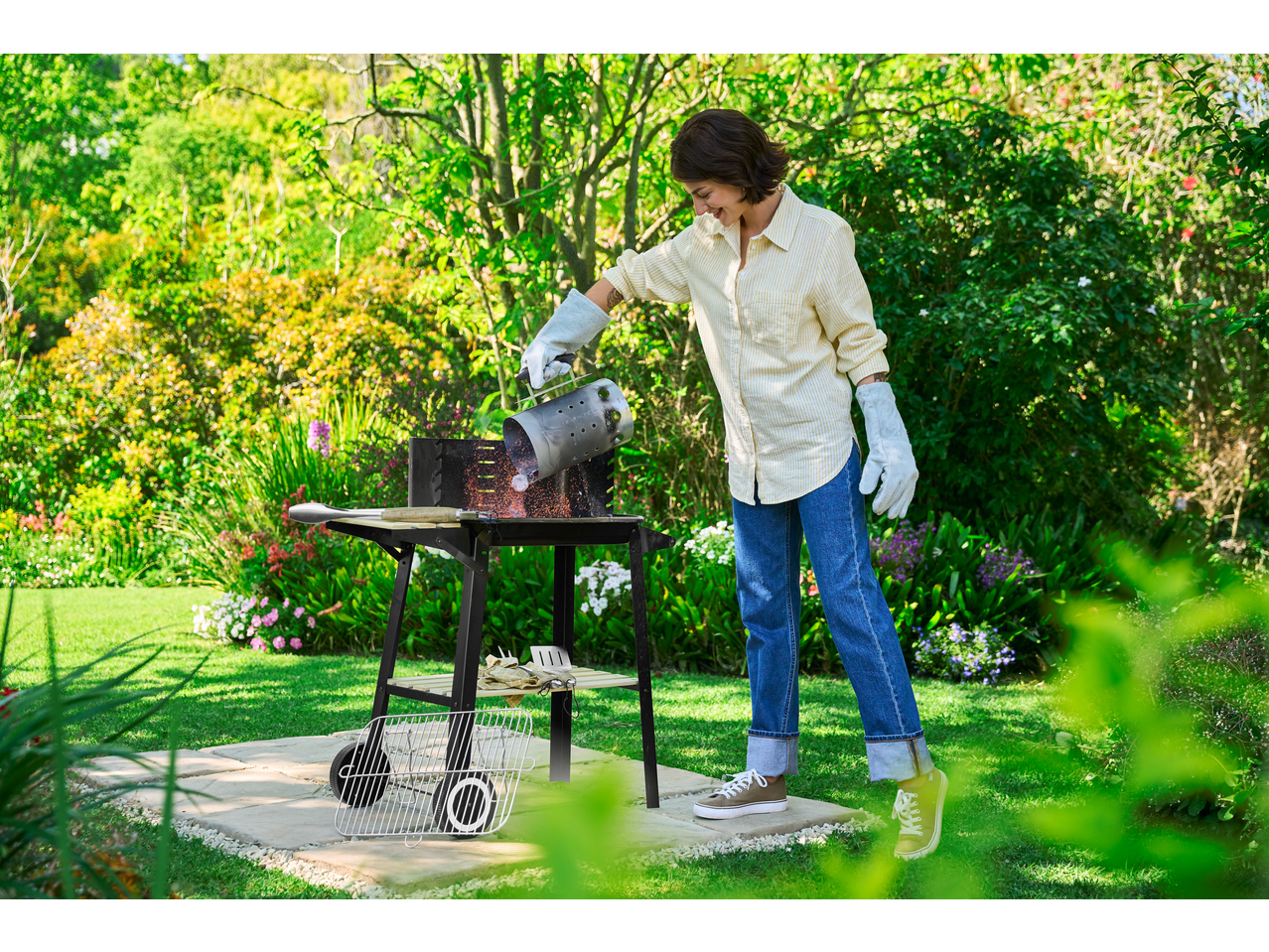 Femeie în mănuși turnând cărbuni dintr-un coș de aprindere în grătar, în grădină.