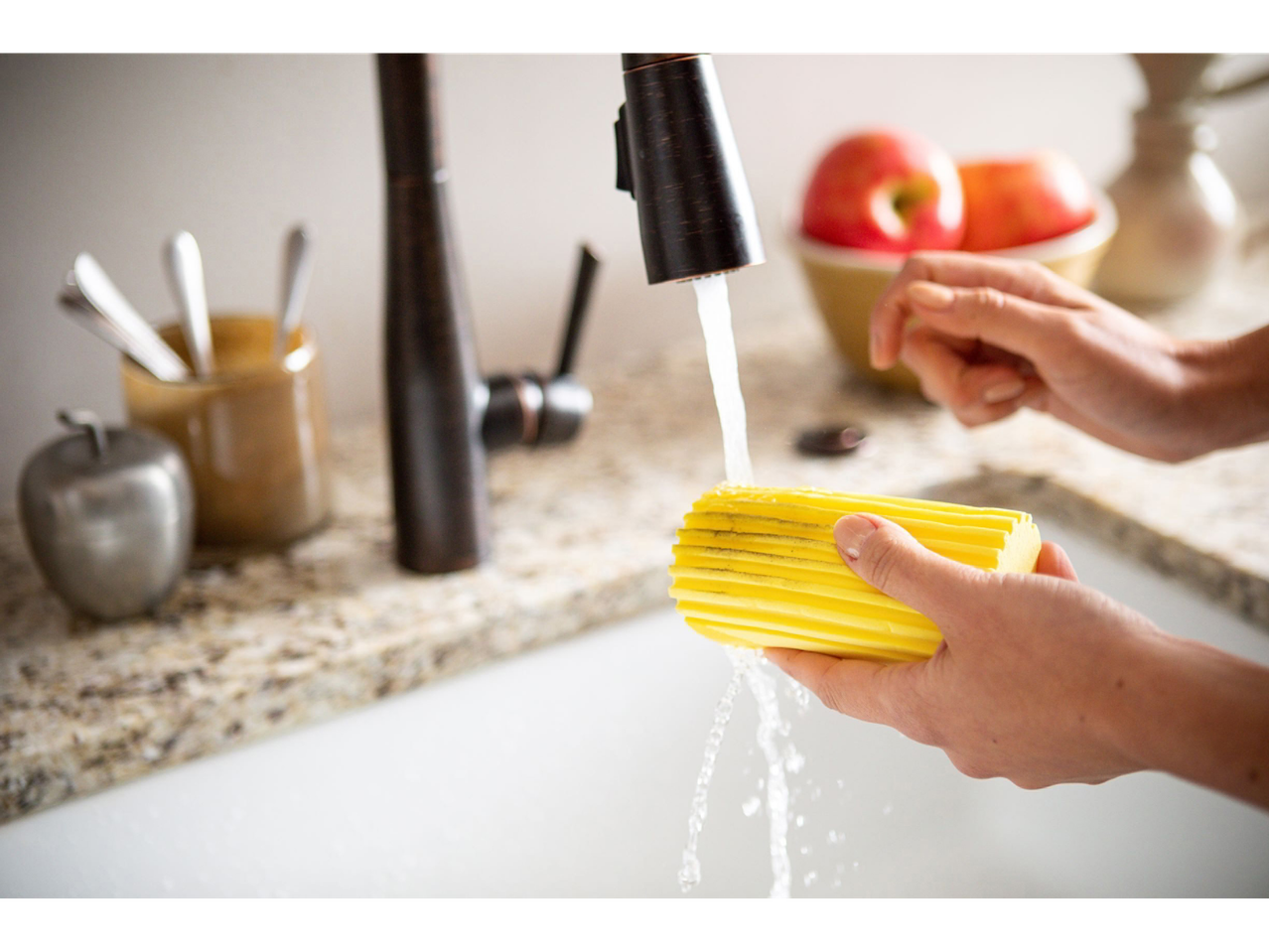 Een hand knijpt een Scrub Daddy spons uit onder stromend water.