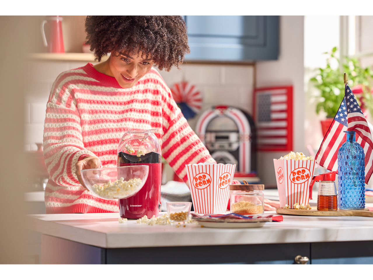 Žena sa Silvercrest® aparatom za kokice i zdjelama kokica u kuhinji s američkom tematikom.