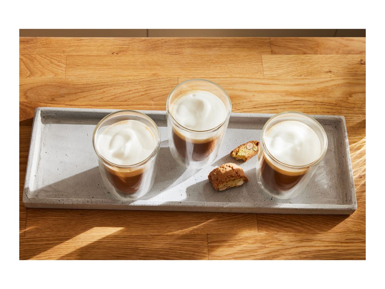 Three glasses of coffee with milk foam and biscotti on a concrete tray.