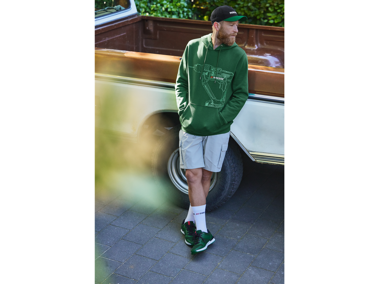 Un bărbat cu un hanorac verde Parkside și adidași verzi stă lângă o camionetă.