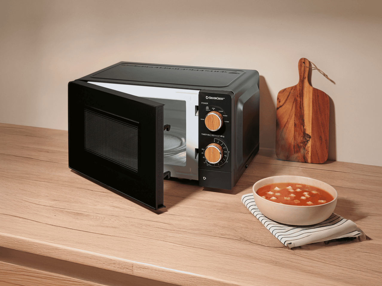 Mikrowelle mit offener Tür, daneben eine Schale Suppe und ein Schneidebrett auf einer Holztheke.