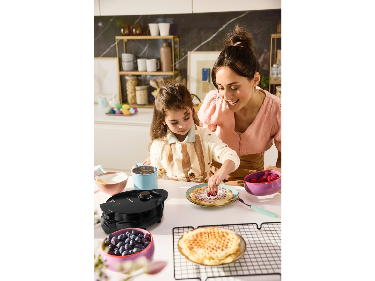 Mother and child making waffles with a waffle maker, topped with berries and powdered sugar.